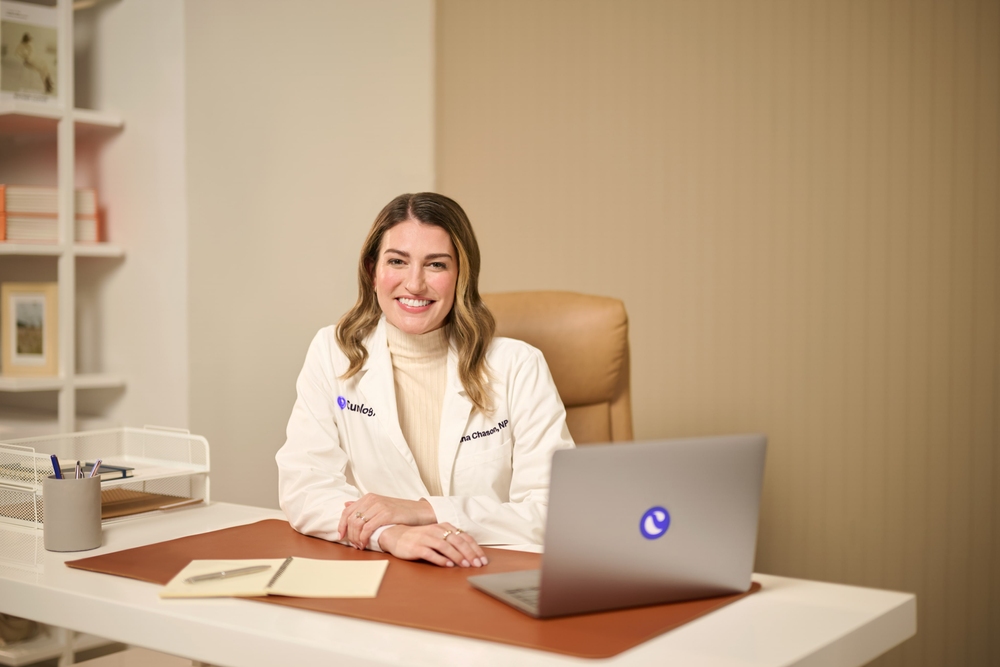 Blonde woman in a white lab coat and beige turtleneck, smiling, sitting behind a desk on a leather chair with a computer.