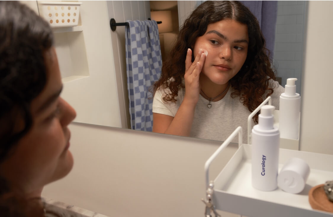 Young brunette woman with curly hair, looking in the bathroom mirror while applying Curology Custom Formula, with the uncapped open bottle on a white shelf next to her.